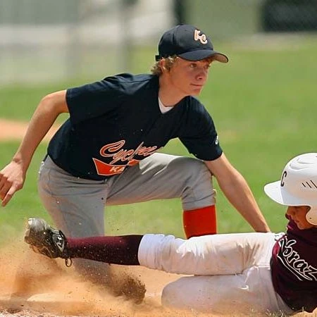 Two players playing baseball. One player is in a base position, kneeling down to catch a low throw. The other is in process of sliding into the base, kicking up a cloud of dirt with him.