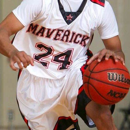 A basketball player dribbles a basketball. The player is dressed in a white uniform white navy blue and orange details. The player's jersey has written across the front the name, Maverick and the number 24.